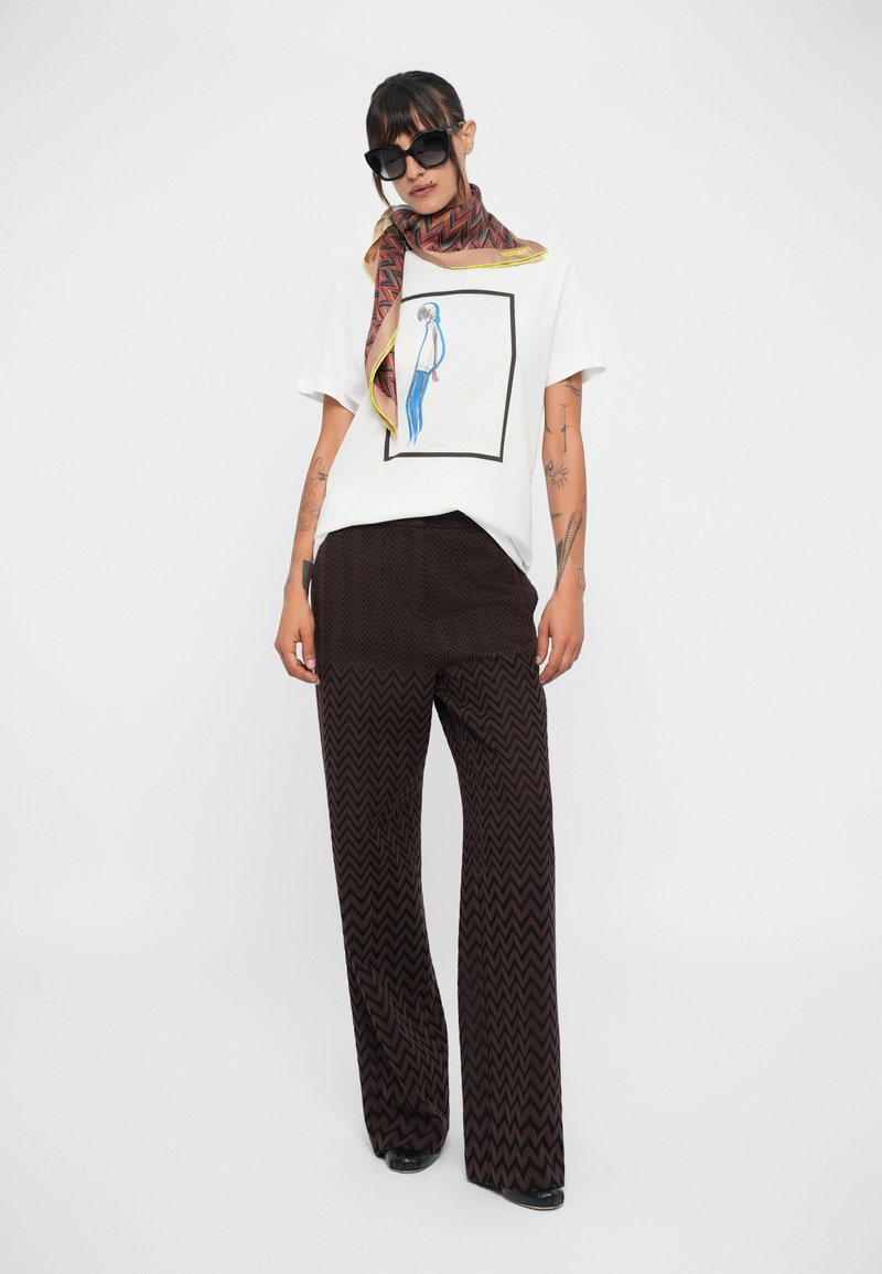 Donna che indossa occhiali da sole neri, una sciarpa rossa a fantasia, una t-shirt bianca con grafica e pantaloni larghi a zigzag neri, in piedi su uno sfondo bianco.