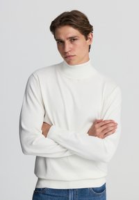 Maglione bianco a collo alto, in tessuto a maglia, con polsini e orlo a coste, design aderente. Modella con le braccia incrociate e indossante jeans in denim blu.