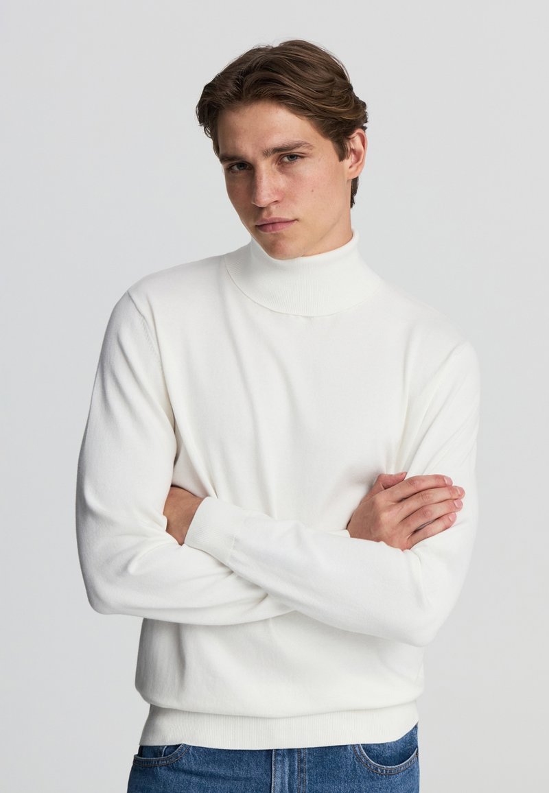 Maglione bianco a collo alto, in tessuto a maglia, con polsini e orlo a coste, design aderente. Modella con le braccia incrociate e indossante jeans in denim blu.