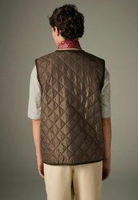 Brown quilted vest with a diamond pattern, featuring a high neckline and contrasting trim, worn over a striped shirt. Red bandana visible.