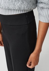 Zwarte op maat gemaakte broek met een gladde textuur, voorzien van een zijrits en een hoge tailleband, gecombineerd met een grijze, gestructureerde gebreide trui.