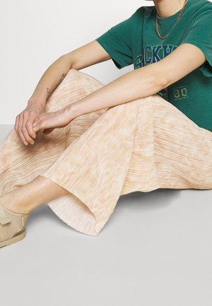 Osoba siedząca po turecku, ubrana w zieloną koszulkę z nadrukiem, beżowe szerokie spodnie plisowane oraz beżowe sandały, z złotym łańcuszkiem na szyi.