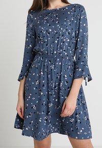 Sininen mekko, jossa on leikkisä täpläkuvio pinkillä, mustalla ja valkoisella. Mekossa on pitkät hihat, joissa on levenevät hihansuut ja elastinen vyötärö.