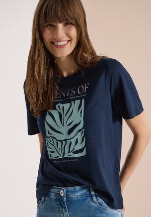 Femme souriante aux longs cheveux bruns portant un t-shirt bleu marine avec un grand motif de feuille abstraite vert clair et un jean bleu.