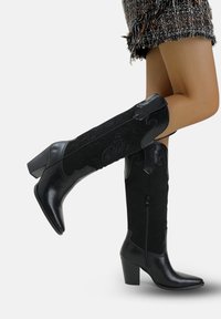 Botas negras hasta la rodilla que combinan cuero liso y ante texturizado, con punta afilada, tacón cuadrado y detalles decorativos en relieve.