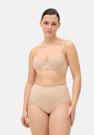 Mujer de cabello oscuro con un sujetador acolchado beige y ropa interior de talle alto sin costuras, posando contra un fondo claro y liso.