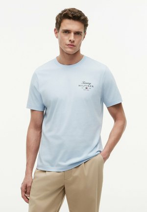 Mężczyzna w jasnoniebieskiej koszulce Tommy Hilfiger i beżowych spodniach, stojący z jedną ręką w kieszeni na białym tle.