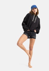 Black pullover hoodie with a textured knit and white drawstrings, paired with distressed black denim shorts. Blue cap with logo accents.
