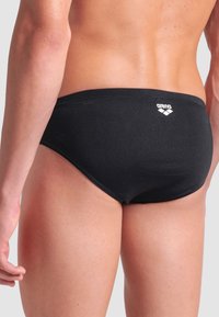 Nahaufnahme einer Person, die schwarze Arena-Schwimmbrieftaschen trägt, die den unteren Rücken, die Hüften und die Oberschenkel gegen einen schlichten Hintergrund zeigt.