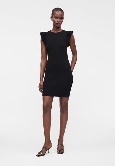 Femme portant une robe noire sans manches ajustée avec des volants aux épaules et des sandales à talons noires ouvertes à l'avant, debout devant un fond uni.