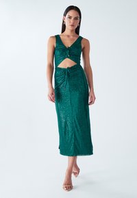 Smaragdinės žalios spalvos suknelė su blizgančiomis akutėmis, aptempta silueto forma, pažemintu iškirptės dekoltė, iškirptu liemens viduriu ir raukinių detalėmis. Rinktinė apranga baigiama plokščiais sandalais.