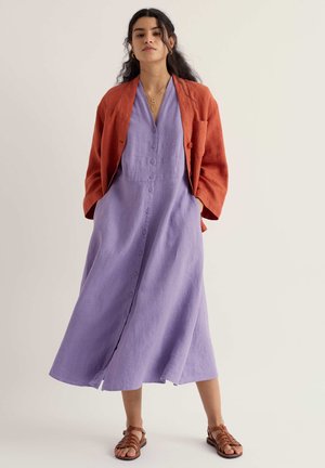 Naine kannab nuppudega avatud lilla midi-kleiti taskutega, rust oranži jope on õlgadel rippumas, jalas pruunid sandaalid ja kaelas kuldne kaelakee.