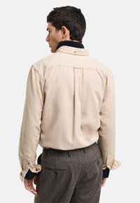 Camisa beige de botones con pliegue en la espalda y mangas enrolladas, combinada con pantalones marrones texturizados. La tela parece suave con un acabado sutil.