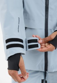 Himmelblaues wasserdichtes Jackett mit schwarzen Klettverschlüssen an den Ärmeln und einem schwarzen Reißverschluss. Verfügt über ein strukturiertes Gewebe und ein "W-PRO 10000"-Etikett.