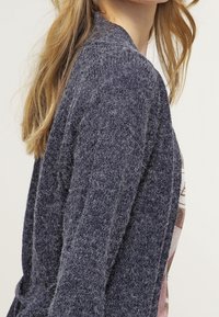 Mörkblå stickad cardigan med strukturerad finish, öppen front och lång ärm, lagd över en ljusfärgad grafisk t-skjorta.