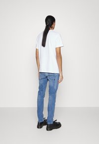 Camiseta blanca de algodón con mangas cortas, combinada con jeans de mezclilla azul claro. Zapatillas negras de suela gruesa. Vista desde la parte de atrás con un fondo liso.