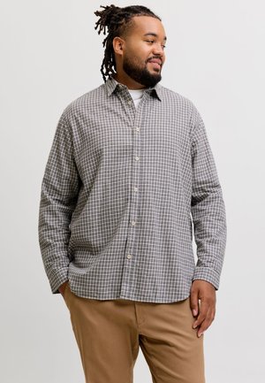 Homme avec des dreadlocks portant une chemise grise à carreaux boutonnée et un pantalon beige, regardant sur le côté les mains dans les poches sur un fond uni.