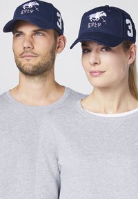 Marineblaues Cap mit gesticktem Polo-Spieler-Logo und der Nummer 3. Kombiniert mit grauen Sweatshirts, die einen Rundhalsausschnitt und eine subtile Textur aufweisen.