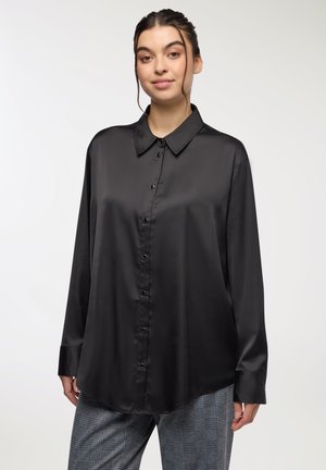 Camicia nera in satin con bottoni, maniche lunghe e colletto a punta; presenta una texture liscia e una finitura lucida, indossata in modo morbido.