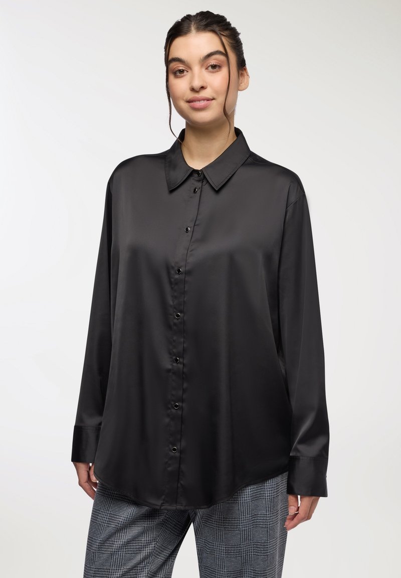 Camicia nera in satin con bottoni, maniche lunghe e colletto a punta; presenta una texture liscia e una finitura lucida, indossata in modo morbido.