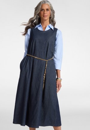 Frau mit grauen Haaren, die ein ärmelloses, dunkelblaues Jeanskleid über einem hellblauen Hemd trägt, accessoriert mit einem goldenen Kettengürtel und Armbändern.