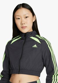 Junge Frau mit langen dunklen Haaren trägt eine schwarze, kurz geschnittene Adidas-Jacke mit neongrünen Streifen und Logo, schaut nach vorne vor einem weißen Hintergrund.