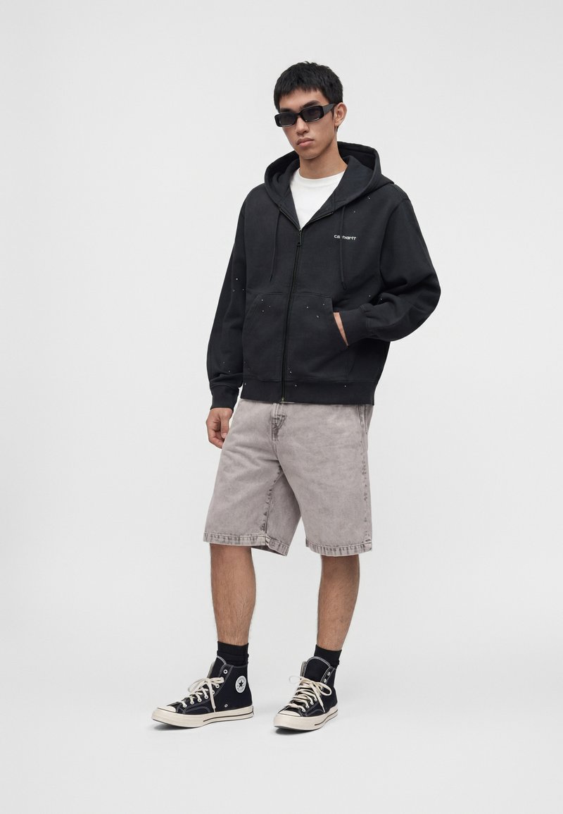 Homme portant un sweat à capuche noir zippé, un short gris clair jusqu'aux genoux, des baskets montantes noires, des chaussettes noires et des lunettes de soleil noires, debout devant un fond uni.