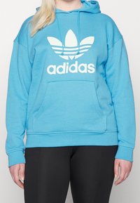 Blå bomullshoodie med en stor vit Adidas-logotyp och grafiken med tre ränder. Har en framficka och justerbar dragsko i huvan.