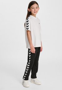 T-shirt blanc avec des rayures en chevron noires sur les manches, associé à un pantalon noir avec des rayures latérales assorties. Porté avec des baskets blanches.