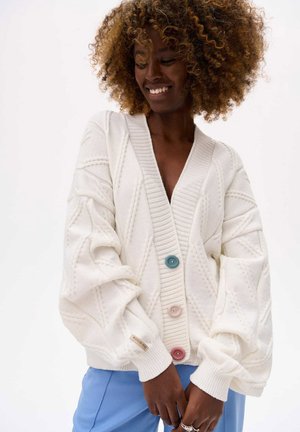 White knitted cardigan with a v-neck, featuring a diamond pattern and three buttons in blue, pink, and white. Textured sleeves and cuffs.