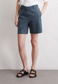Dark teal cotton shorts with a straight cut, featuring a front zipper and side pockets, paired with black slide sandals with gold accents.