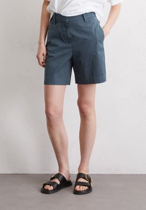 Shorts en coton bleu foncé avec une coupe droite, dotés d'une fermeture éclair à l'avant et de poches latérales, associés à des sandales noires à glissière avec des accents dorés.