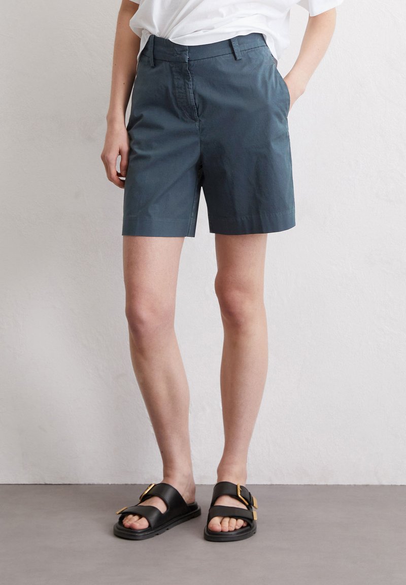 Dark teal cotton shorts with a straight cut, featuring a front zipper and side pockets, paired with black slide sandals with gold accents.