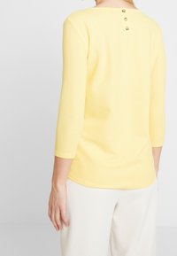 Žena s bledě žlutým texturovaným topem s třičtvrtinovými rukávy a dvěma malými knoflíky na horní části zad a bílými kalhotami.