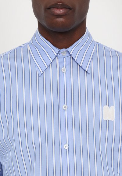 Camisa de rayas azul claro con cierre de botones blancos, que presenta un cuello puntiagudo y un logo blanco bordado en el lado izquierdo.