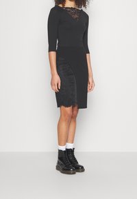 Svart bodycon-klänning med spetsdetaljer vid halsringningen, trekvartsärmar och strukturerad blommig spetsaccent på en sidoflikpanel. Svarta kängor.