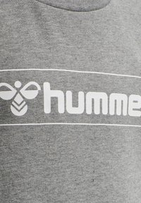 Maglietta in cotone grigia con un design grafico bianco che presenta un'icona di un'ape e il testo "humme". Collo tondo, maniche corte, texture liscia.