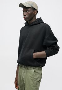 Μαύρη φαρδιά φούτερ με καδένα μπροστά, συνδυασμένη με ελαιοπράσινα cargo παντελόνια. Το μοντέλο φορά επίσης ένα ανοιχτό γκρι καπέλο.