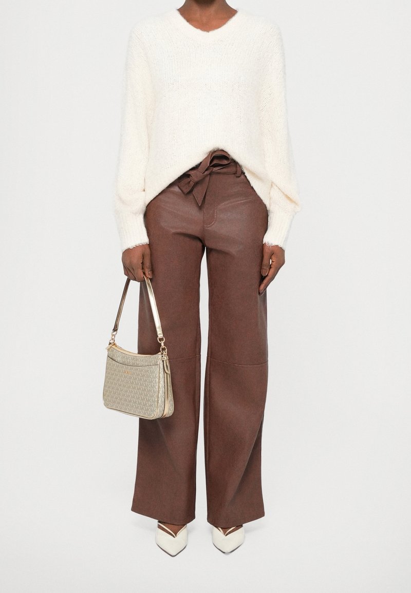 Personne portant un pull blanc, un pantalon marron à jambes larges avec une ceinture nouée, des chaussures blanches pointues, et tenant un sac à main beige à motif dans une main.