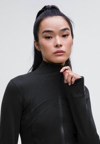 Haut ajusté noir à col montant avec fermeture éclair, présentant un panneau texturé et des détails de coutures. Tissu lisse avec une texture extensible pour le confort.