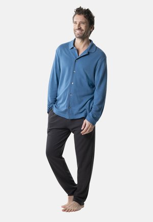 Chemise à manches longues bleue avec boutons en tissu texturé, associée à un pantalon noir, présentée sur un homme debout pieds nus.