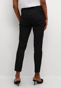 Pantalons noirs ajustés avec une texture lisse, dotés d'une taille mi-haute et de deux poches arrière. Portés avec des mules à talons ouverts.