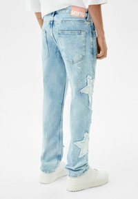Persona que lleva puestos unos pantalones de mezclilla azul claro con parches en forma de estrella y zapatillas blancas, de pie contra un fondo blanco liso.