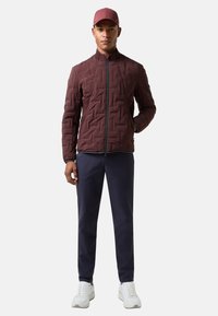 Bordeauxrote gesteppte Jacke mit hohem Kragen und Reißverschluss, kombiniert mit marineblauer Hose und weißen Sneakern. Der Stoff hat ein gestepptes Muster.