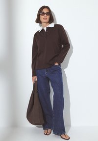 Rudas ilgomis rankovėmis polo marškinėliai su baltu apykakle, derinami su mėlynomis plačiomis kelnėmis ir rudų spalvų rankine. Modelis stovi prieš baltą foną.