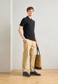 Černé polo tričko, béžové kalhoty, černé boty se šedou podrážkou, držící hnědou tašku na rameno. Hladká tkanina a upravený střih. Přírodní dřevěná podlaha.