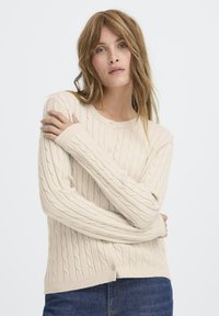Beige strikket cardigan med kabler, rund halsudskæring og knaplukning. Har lange ærmer samt ribbede manchetter og kant.