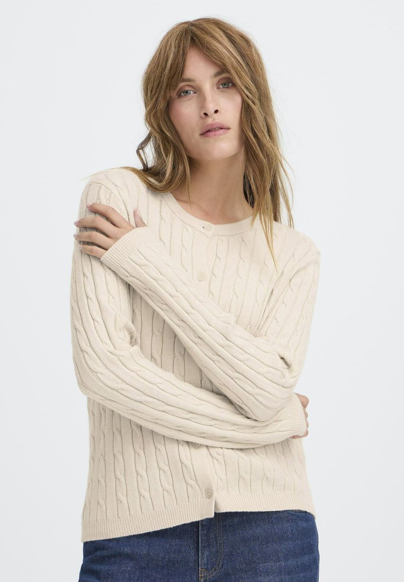 Beige strikket cardigan med kabler, rund halsudskæring og knaplukning. Har lange ærmer samt ribbede manchetter og kant.