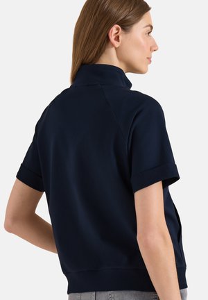 Femme aux cheveux châtain clair portant un haut bleu marine à manches courtes avec un col montant, vue de dos et de côté sur fond blanc.