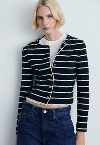 Skraćeni tamnoplavi kardigan s bijelim prugama, zlatnim gumbima i okruglim izrezom, nosi se preko bijele majice i uparen je s tamnim trapericama.